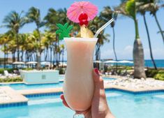 a person holding up a drink in front of a swimming pool with palm trees and umbrellas