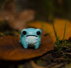 a small blue frog toy sitting on top of a leaf covered ground with grass in the background