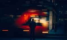 a man standing on the sidewalk in front of a red bus with blurry lights