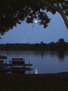 the moon shines brightly in the night sky over a lake with benches and an umbrella