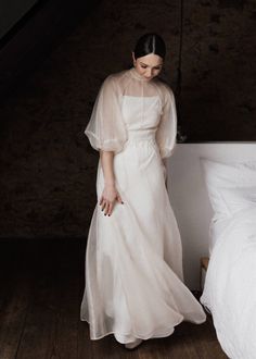 a woman in a white dress standing next to a bed and looking down at the floor