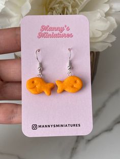 a pair of tiny yellow fish earrings sitting on top of a pink card next to a white flower