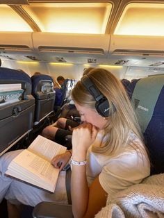 a woman sitting in an airplane reading a book and listening to headphones on her ear