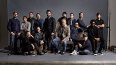 a group of men posing for a photo in front of a gray background with one man sitting on a chair