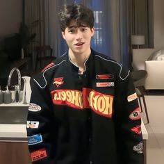 a young man wearing a black jacket with hot wheels on the front and chest, standing in a kitchen