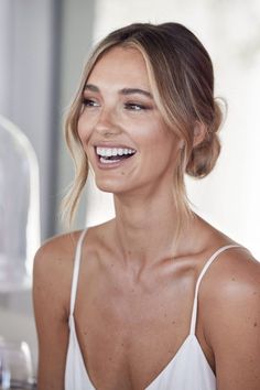 a woman smiling and wearing a white tank top with her hair in a low bun