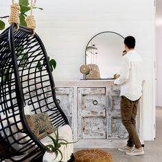 a man standing in front of a chair next to a dresser and table with a mirror on it