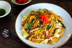 a white plate topped with noodles and veggies next to chopsticks on a wooden table