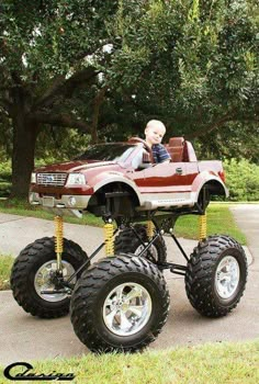 a small child in a toy truck with big wheels on the street next to trees