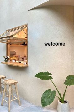 a welcome sign on the side of a white wall next to two stools and a potted plant