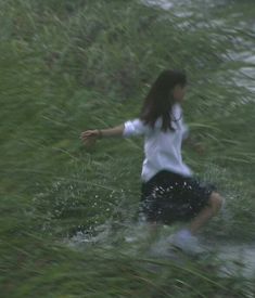 a woman in white shirt and black shorts walking through water with green grass behind her