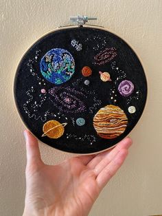 a hand is holding up a black embroidery project with planets and stars in the sky