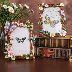 two frames with flowers and butterflies are sitting on a table next to books that have been placed in front of them