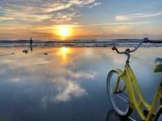 a yellow bike parked on top of a wet beach next to the ocean at sunset