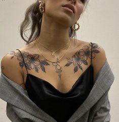 a woman with tattoos on her chest wearing a black dress and gold hoop earrings, standing in front of a white wall