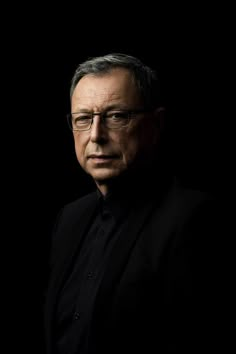 an older man wearing glasses and a black shirt is looking at the camera while standing in front of a dark background