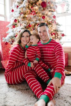 a man and woman in matching pajamas are sitting next to a christmas tree while holding their child