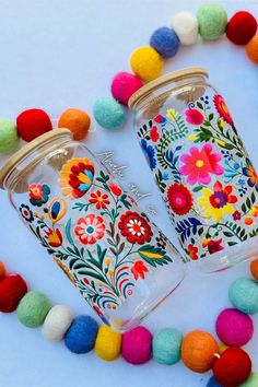 two glass jars with colorful flowers and pom - poms are on the table