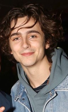 a close up of a person wearing a denim jacket and smiling at the camera with one hand on his hip