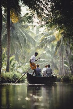 three people in a boat on a river surrounded by palm trees and greenery, with one man holding a guitar