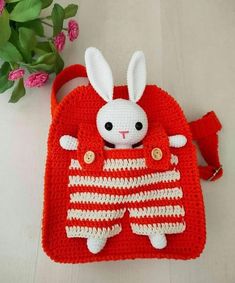 a crocheted red purse with a white rabbit in the pocket and flowers behind it