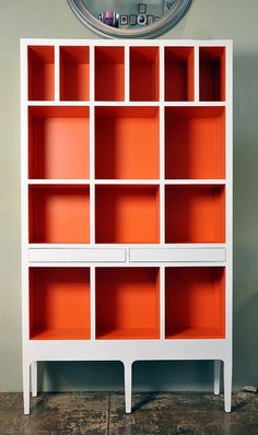 a white bookcase with red shelves and a clock on the wall behind it in a room