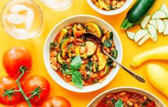 three bowls of food on a yellow table with vegetables and wine glasses next to them