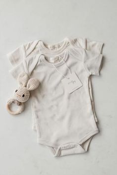 Two cream-colored baby bodysuits, one placed on top of the other, accompanied by a crocheted bunny rattle with a wooden ring, laid out on a light marble surface. Cute Wardrobe, Baby Wardrobe, Bodysuit Designs, Easy Dressing