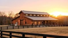 a barn with a fence in the foreground and trees in the background at sunset