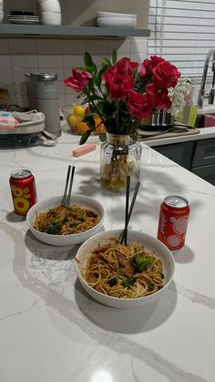 two bowls of noodles and flowers on a kitchen counter with cans of soda, can openers and utensils