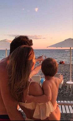 a man and woman holding a baby on the beach at sunset with umbrellas in the background