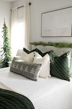 a white bed topped with lots of pillows next to a christmas tree in a bedroom