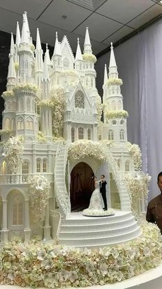 a wedding cake that looks like a castle with white flowers on the side and a bride and groom standing in front