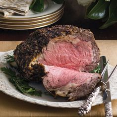 a large piece of meat on a plate next to a knife and fork with some flowers in the background