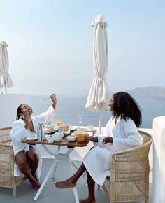 two women sitting at a table with food and drinks in front of an ocean view