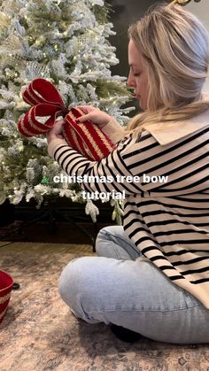 a woman sitting in front of a christmas tree tying her red ribbon around the bow