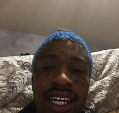 a man with blue hair and tattoos on his face smiles at the camera while lying in bed