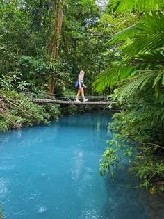 a woman walking across a bridge over a river in the jungle with blue water and trees