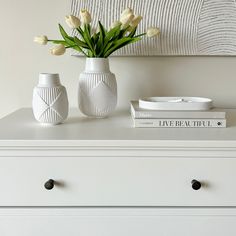 two white vases sitting on top of a dresser next to a book and flowers