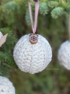 three ornaments hanging from a tree with pine cones and leaves in the foreground, one white ornament has a brown button on it's end