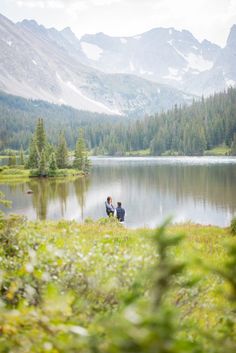 two people sitting on the shore of a lake with mountains in the backgroud