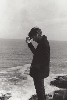 a man standing on top of a cliff next to the ocean holding a cell phone