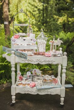 a white table topped with lots of desserts next to lush green trees and bushes