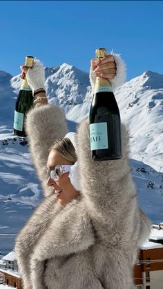 a woman holding two bottles of champagne in front of snow covered mountains and blue sky