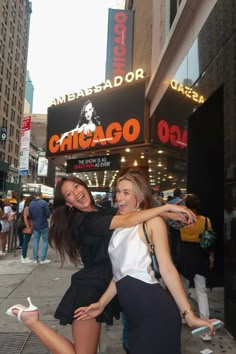 two beautiful women standing next to each other in front of a theater sign and buildings