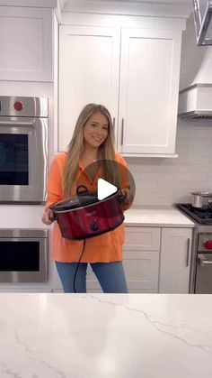 a woman standing in a kitchen holding a red bag with the handles down and an oven behind her