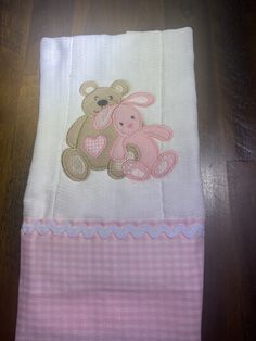 a pink and white blanket with a teddy bear embroidered on the side, sitting on a wooden floor