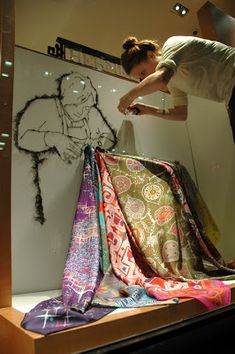 a woman standing on top of a display case filled with scarves and cloths