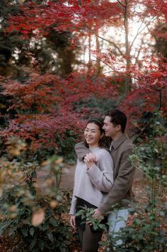 a man and woman standing next to each other in front of trees with red leaves