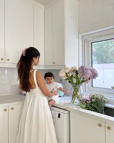 a woman in a white dress standing at a kitchen counter with a baby on her lap